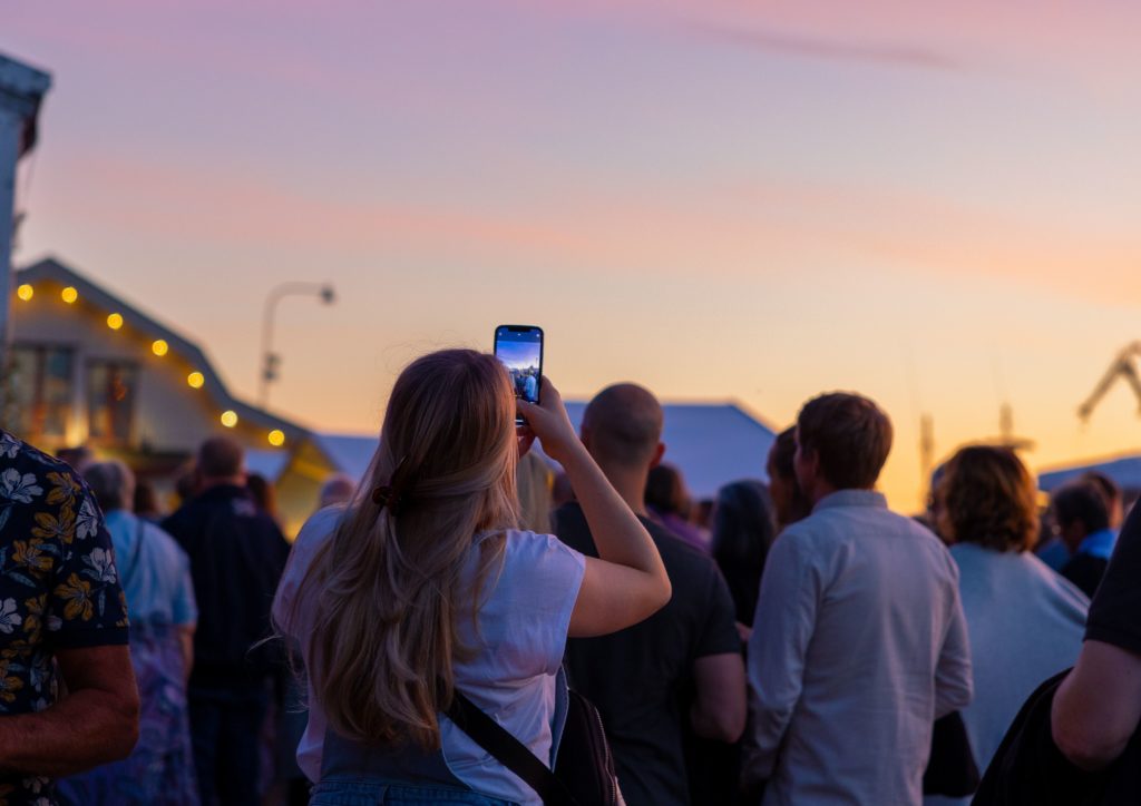 Bild på mnniskor i solnedgång framför scen som tar bilder med sina mobiler.